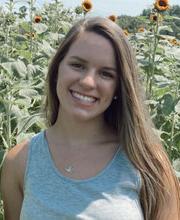 Olivia Dean, '22, smiling with long straight light brown hair and wearing a light blue tank top with sunflowers in the background